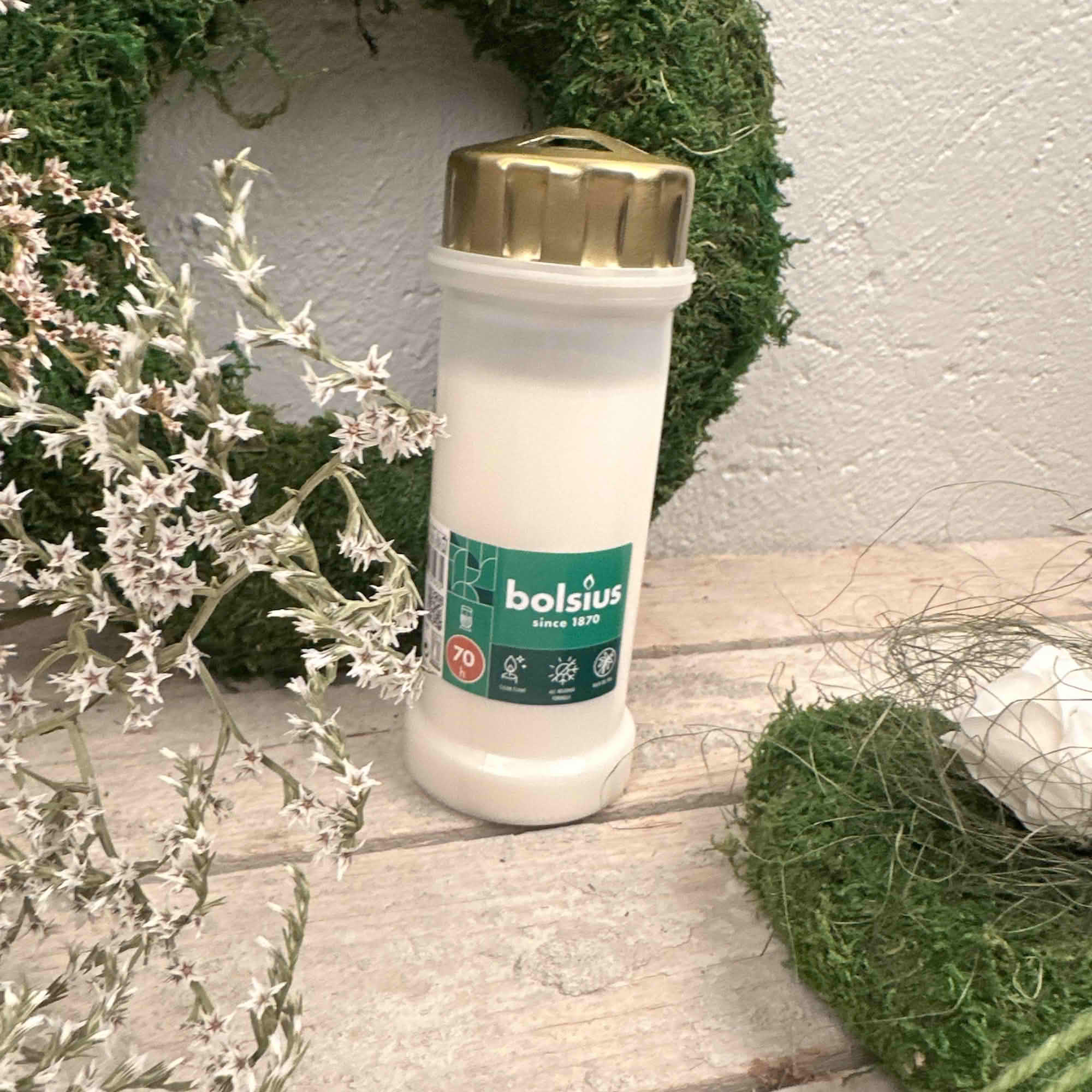 Vitt Bolsius gravljus med guldlock framför en grön mosskrans